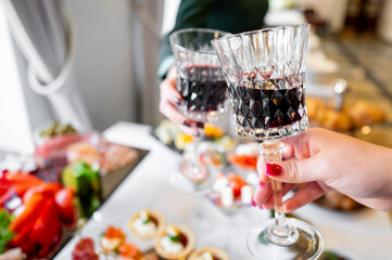 Two hands holding glasses of red wine, toasting over a blurred background of a food buffet. The image captures a celebratory moment, perfect for themes of dining, celebration, and social gatherings.