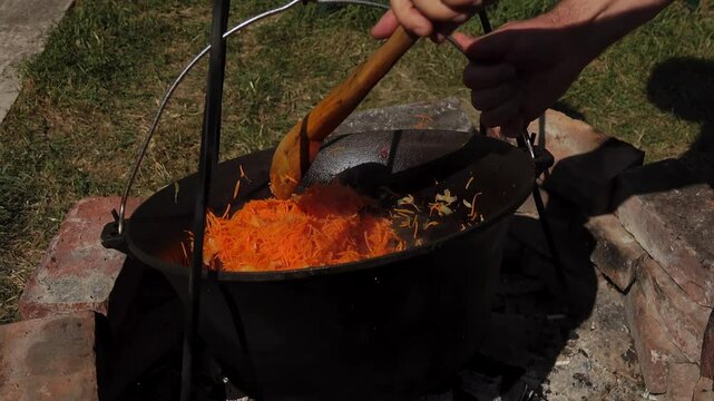 A traditional Romanian cooking scene showing onions being saut&eacute;ed in a cauldron over a wood fire, a key step in making authentic zacusca. The rustic outdoor setup highlights the time-honored methods.