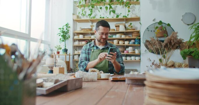 Camera focus on happy Caucasian male texting with someone on smartphone. Communicating remotely with friends or family with help of mobile device. In background shelves with completer pottery projects
