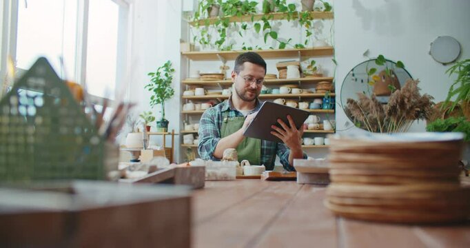 Positive Caucasian potter working in studio. Holding tablet device horizontally. Looking at screen while typing message. Swiping on screen with fingers. Searching for ideas online.