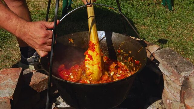 A traditional Romanian cooking moment where tomato puree is poured into a cauldron over a wood fire. This rustic outdoor scene captures the making of authentic zacusca, a flavorful spread