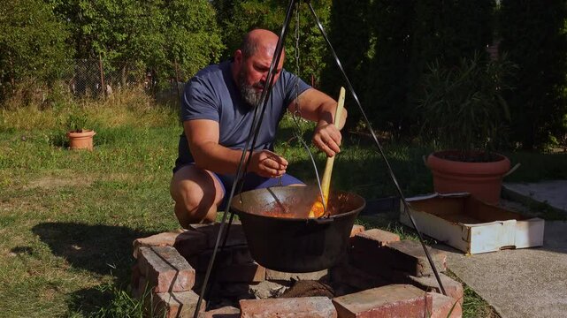 A traditional Romanian cooking moment where tomato puree is poured into a cauldron over a wood fire. This rustic outdoor scene captures the making of authentic zacusca, a flavorful spread.