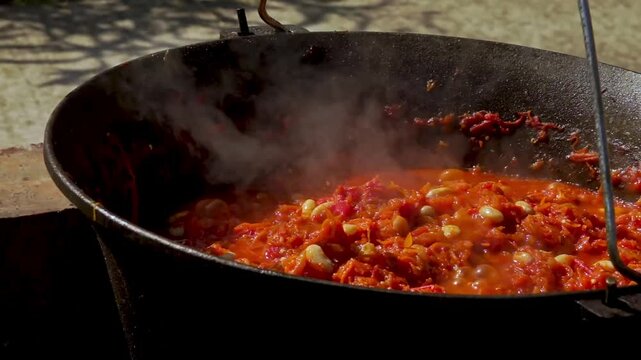 A traditional Romanian cooking moment where tomato puree is poured into a cauldron over a wood fire. This rustic outdoor scene captures the making of authentic zacusca, a flavorful spread