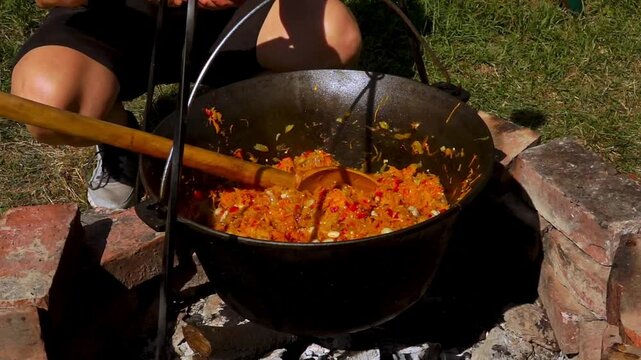 A traditional Romanian cooking moment where tomato puree is poured into a cauldron over a wood fire. This rustic outdoor scene captures the making of authentic zacusca, a flavorful spread