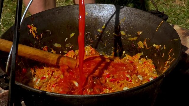 A traditional Romanian cooking moment where tomato puree is poured into a cauldron over a wood fire. This rustic outdoor scene captures the making of authentic zacusca, a flavorful spread