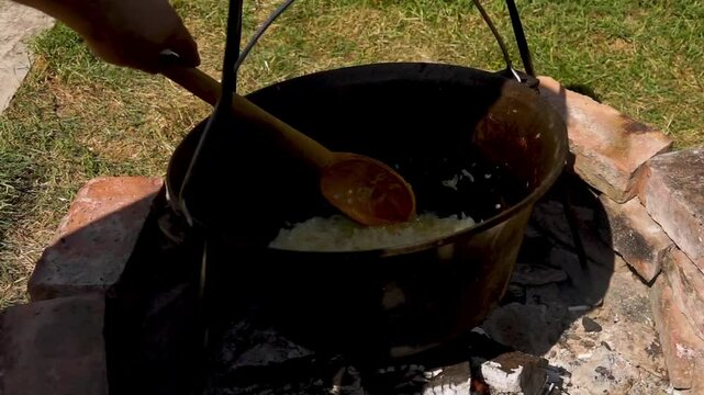 A traditional Romanian cooking scene showing onions being saut&eacute;ed in a cauldron over a wood fire, a key step in making authentic zacusca. The rustic outdoor setup highlights the time-honored methods.