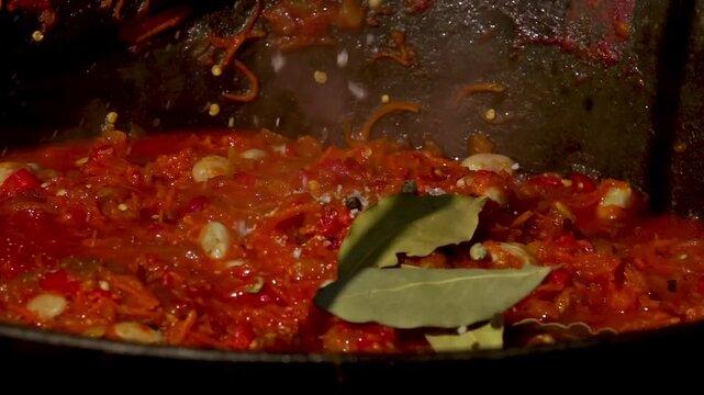 A traditional Romanian cooking moment where spices are added to a simmering cauldron of zacusca over a wood fire. This rustic outdoor scene captures the aromatic blend of flavors.