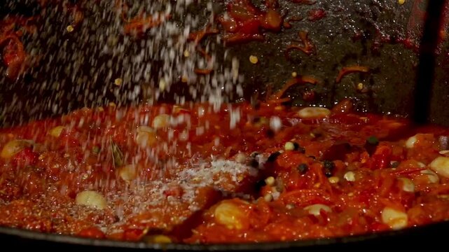 A traditional Romanian cooking moment where spices are added to a simmering cauldron of zacusca over a wood fire. This rustic outdoor scene captures the aromatic blend of flavors.