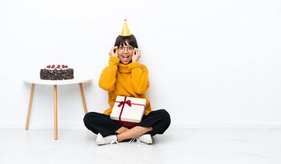 Young mixed race woman holding a gift sitting on the floor isolated on white background with glasses and surprised