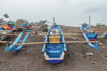 This photo shows a row of colorful fishing boats anchored by the shore. The boats are dominated by blue and orange colors, with names written on their hulls. The masts of the sails can be seen looming