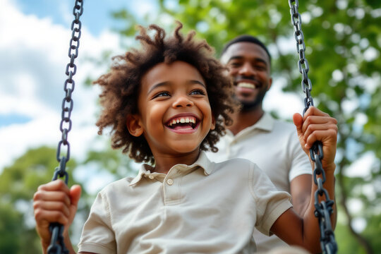 A joyful African American father stands behind his young son who is swinging on a swing in a lush green park. - Powered by Adobe