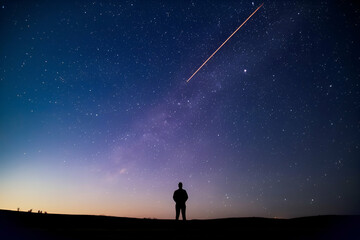 Fototapeta premium A silhouette of a person gazes at a starlit sky, with a meteor streaking above, creating a serene and awe-inspiring cosmic scene.