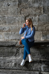 Beautiful young woman wearing jeans and jacket sitting on stone wall against background of stone wall of old building talking on phone on autumn day. Break in sightseeing in old town. Bergamo, Italy. 