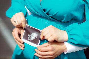 A pregnant woman in the arms of her husband. Summer sunny weather. A couple in love are hugging. Ultrasound picture with the baby's gender in the mother's hands.