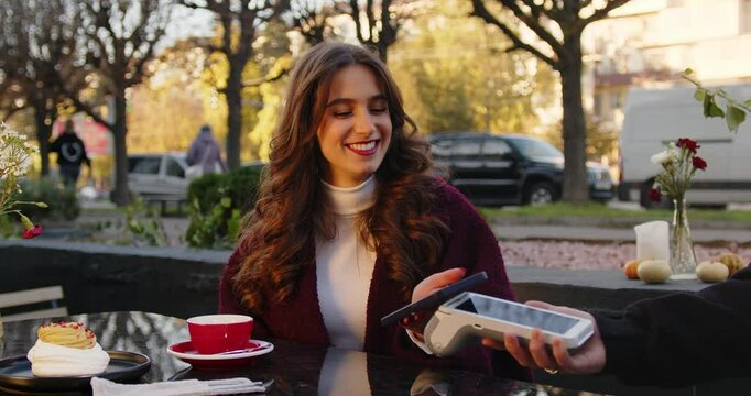 Cheerful young woman using smartphone to pay for her order in a cafe, smiling, enjoying modern technology. People and money concept.