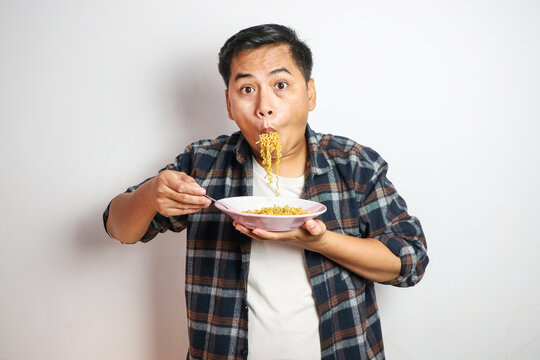 Young handsome trendy asian man eating yummy hot and spicy instant noodle using fork isolated on white background. Asian guy servile end of the month with cheap food.