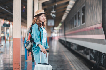 Obraz premium A young woman in casual attire and hat stands with a suitcase on a train station platform, ready for travel.