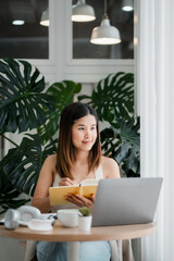 A young woman multitasks by reading a book and working on her laptop in a stylish home office filled with lush green plants.