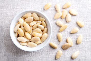 Freshly blanched almonds in a white bowl on linen. Shelled almonds that have been treated with hot water to soften the seed coat, which is then removed to reveal the seed. Fruits of Prunus amygdalus.