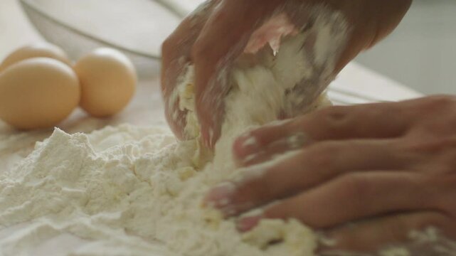 Close-up view of hands kneading mixture of flour and eggs on kitchen table, preparing dough or homemade pasta, with couple of whole eggs and sieve visible in the background