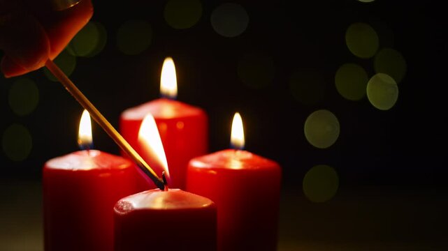 Hand lighting a fourth red candle in the Advent wreath using a match with festive bokeh lights in the background, close up shot. Holiday mood concept.