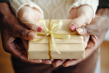 Close-up of four hands holding a small gift box tied with a golden ribbon, highlighting...