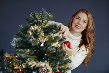 Woman with red hair smiling while decorating Christmas tree with festive ornaments, surrounded by tinsel and pine cones. Wearing cozy sweater in warm setting