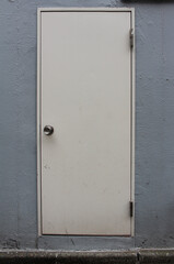 Back door of a building in a street. White door with metal doorknob on grey wall background.