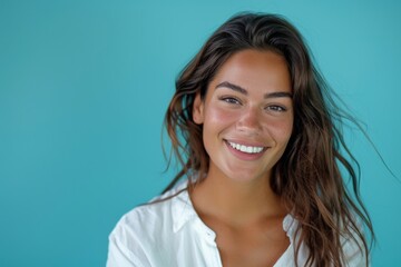 Fototapeta premium Smiling brightly, a Caucasian woman in a white shirt poses confidently against a turquoise backdrop, highlighting her charm and beauty.
