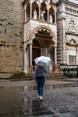 Beautiful blonde woman wearing jeans and jacket holding umbrella walking to basilica in Bergamo, Italy on rainy autumn day. Back view. Santa Maria Maggiore Basilica.