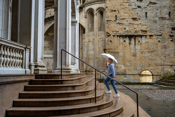 Beautiful young woman wearing jeans and jacket holding umbrella walking up stairs to cathedral in...