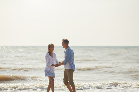 A couple holding hands and smiling at each other while standing in the shallow waves of a beach at sunrise. A romantic and serene moment by the sea.