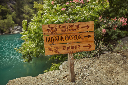 Wooden sign in Goynuk Canyon indicating directions for canyoning and ziplining activities. Surrounded by lush greenery, perfect for nature tourism and outdoor adventure promotions. 