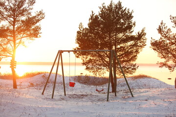 Swing on a snow-covered bank between pine trees against the backdrop of a winter sunset
