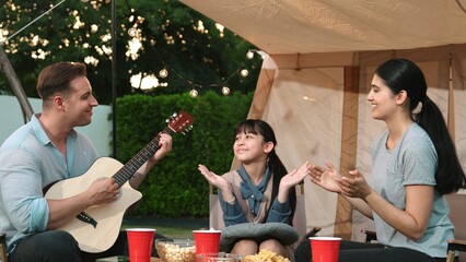 Family together camping. Mother and daughter listening father play guitar. Parent use outdoor camping activity to communicate and spending time with young generation cross generation gap. Divergence.