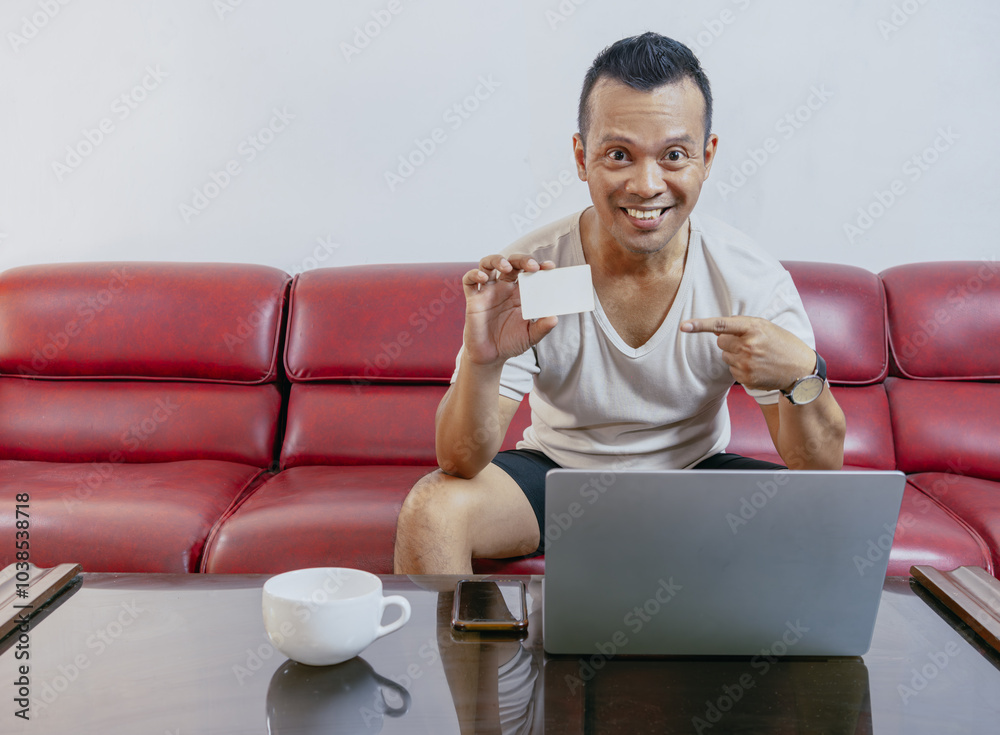 Wall mural asian man smiling happy and showing blank credit card while sitting on the sofa in front of the lapt