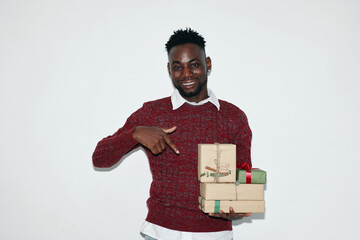 Smiling man holding wrapped gifts and wearing a red sweater against white background, looking at camera. Presenting cheerful expression while pointing at the stack of presents