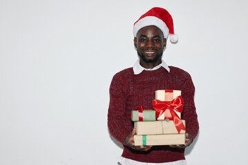 African American man wearing Santa hat while smiling warmly, holding wrapped Christmas presents against white background with joyful expression reflecting holiday cheer