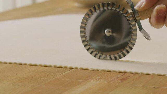 Hand of unrecognizable person cutting dough with fluted pastry wheel cutter on wooden kitchen table. Close-up shot