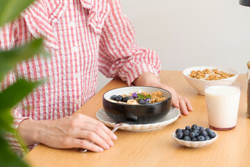 A woman is sitting at a table, eating a healthy breakfast. Granola with Greek yogurt and blueberries