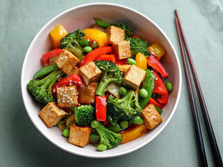 A colorful bowl of vegan stir-fry with tofu, bell peppers, broccoli, and snap peas, accompanied by chopsticks ai.