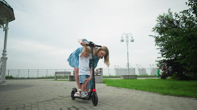 A mother helps her young son stand on a scooter while they are on an interlocked path, she carefully guides him on how to balance, with three women walking in the background