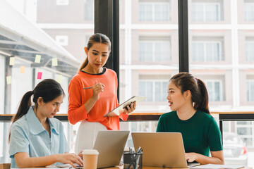 Collaborative meeting with three women discussing ideas in modern office space. One woman is presenting notes while others engage with laptops, showcasing teamwork and productivity