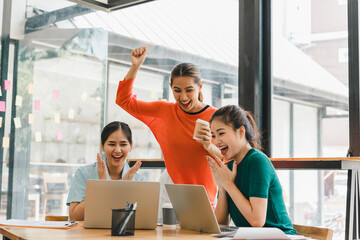 Excited women celebrating success while working on laptops in modern office. Their joyful expressions reflect teamwork and achievement in collaborative environment
