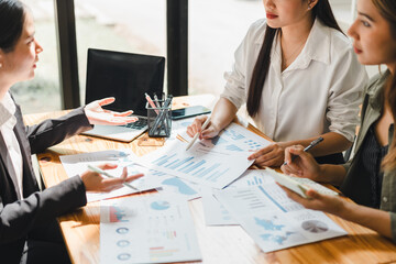Collaborative business meeting with three women discussing charts and graphs, analyzing data on printed reports, and using digital devices. atmosphere is focused and productive