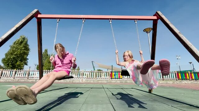 Two children joyfully swing on a playground under a clear blue sky, their feet forward as they enjoy the moment. Family time, sisters.