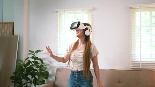 A young woman is sitting on a couch, wearing a virtual reality headset. She raises one hand, seemingly interacting with the virtual environment, with plants