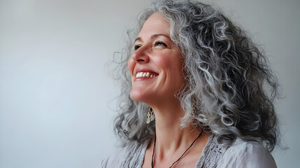 A joyful woman with curly gray hair smiles brightly, embodying happiness and confidence against a simple backdrop