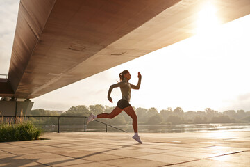 An Energetic Young Woman is Running Swiftly Under a Bridge During a Beautiful Sunrise Scene