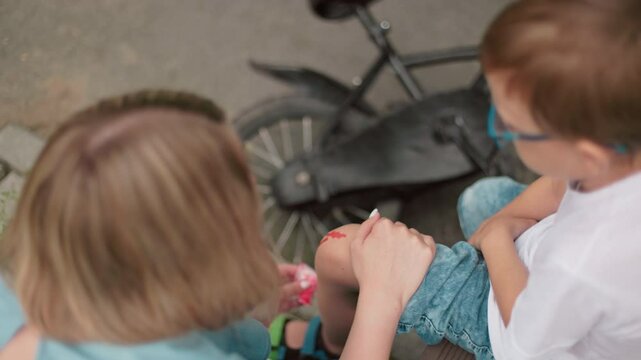 A mother kneels down to clean her son's bleeding leg while his bicycle lies on the ground beside them